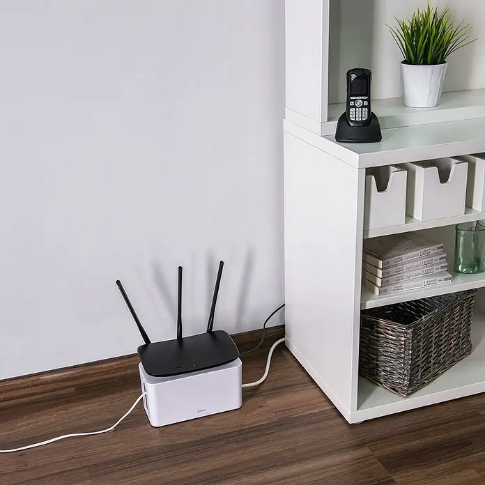 Hama Kabelbox Mini - Weiß - WLAN-Router mit drei Antennen und Kabeln, der auf einem weißen Kasten auf einem Holzboden neben einem weißen Regal steht.