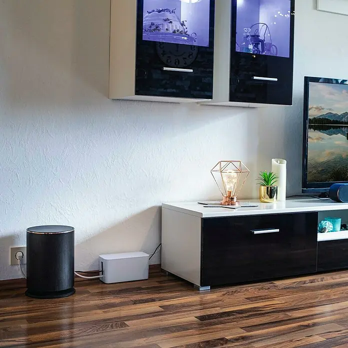 Hama Kabelbox Mini - Weiß - Das Bild zeigt einen schwarzen, zylindrischen Lautsprecher und eine weiße Kabelbox auf einem Holzboden. Daneben steht ein weiß-schwarzes TV-Möbel.