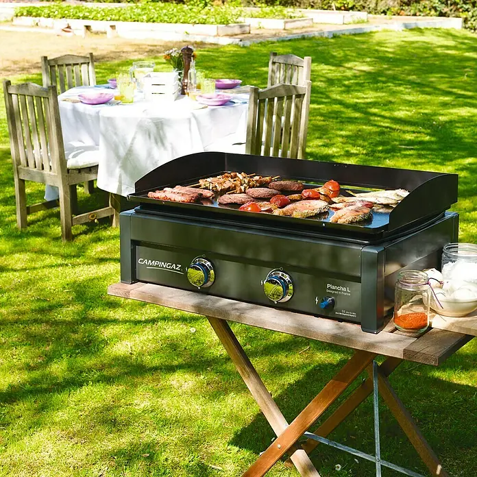 Campingaz Barbacoa de gas L - Superficie de la parrilla (An x Pr): 66 x 42 cm, Número de quemadores: 2 - Una parrilla Campigaz sobre una mesa de madera al aire libre. Se están cocinando varias carnes en la parrilla. Al fondo se puede ver una mesa puesta.