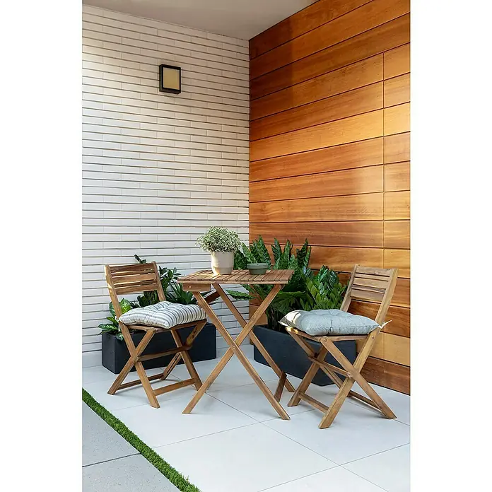 Conjunto de muebles de balcón Bristol - 3 pzs., Madera de acacia, Marrón - Esta imagen muestra un encantador conjunto de bistró al aire libre con una mesa de madera y dos sillas plegables con cojines cómodos, rodeado de exuberante vegetación.