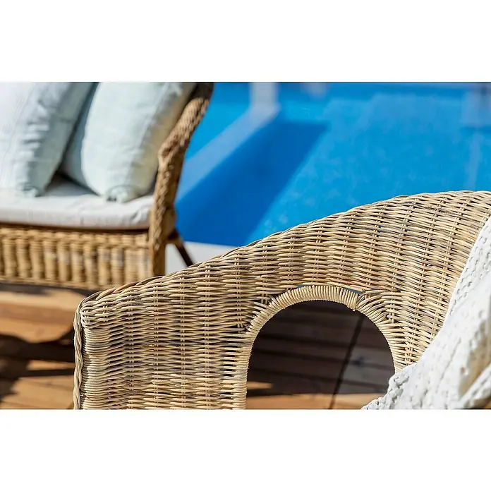 Conjunto de muebles Paula - 4 pzs., Ratán natural, Natural - Primer plano de una silla de ratán marrón claro en el exterior, adornada con almohadas claras y un paño blanco, con vistas a una piscina.