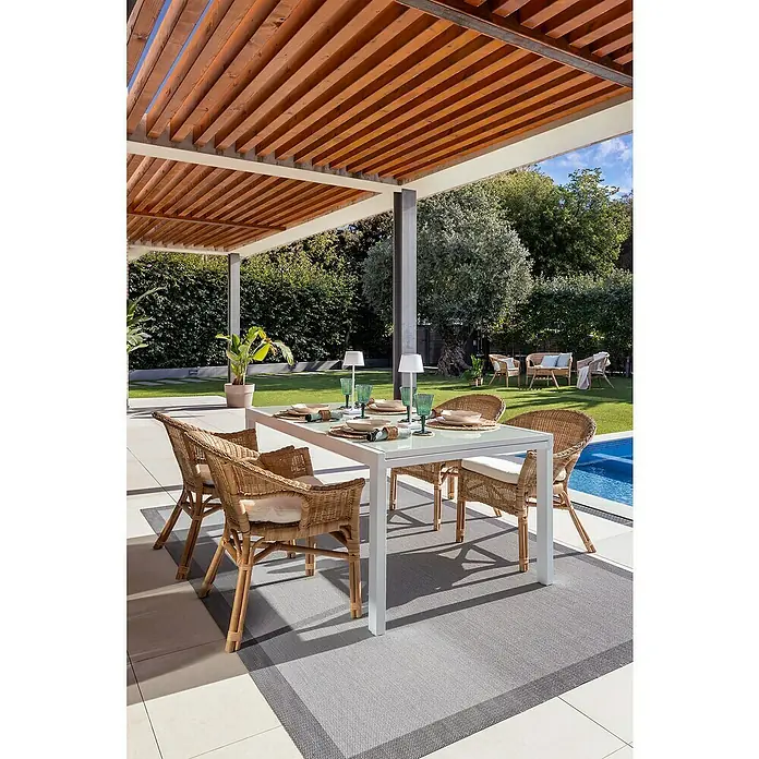 Sunfun Mesa de jardín Petra - L x An x Al: 165 x 100 x 75 cm, Blanco - Imagen de un conjunto de mesa de comedor de ratán con mesa blanca y alfombra de mesa gris al aire libre bajo una pérgola.