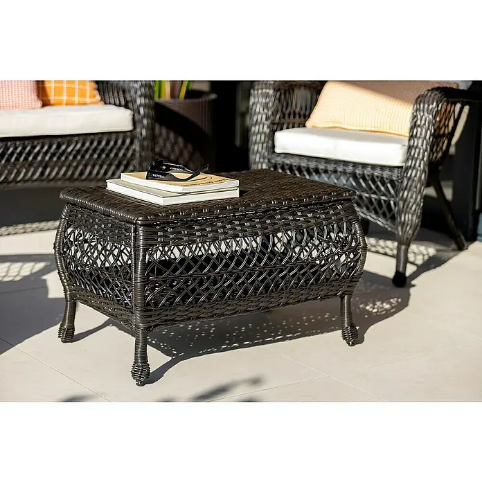Sunfun Conjunto de muebles Savannah - 4 pzs., Ratán sintético/Acero, Marrón oscuro/Beige - Primer plano de una mesa de almacenamiento de ratán marrón con dos libros y gafas de sol encima, con dos sillones al fondo.