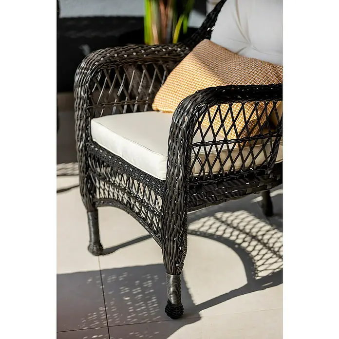 Sunfun Conjunto de muebles Savannah - 4 pzs., Ratán sintético/Acero, Marrón oscuro/Beige - Primer plano de una silla de mimbre negra con cojines blancos y un cojín decorativo amarillo sobre una superficie exterior de baldosas claras.