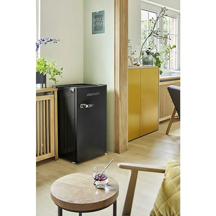 Respekta Vollraumkühlschrank Rico - Nutzinhalt Gesamt: 92 l, 44,7 x 47,4 x 83,1 cm, Schwarz - Dieses Bild zeigt einen schwarzen Retro-Minikühlschrank neben einem Tisch mit Getränk in einem hellen, modernen Raum.