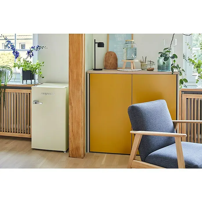 Respekta Vollraumkühlschrank Rico - Nutzinhalt Gesamt: 92 l, 44,7 x 47,4 x 83,1 cm, Cream - Ein stilvolles Interieur mit einem kleinen, cremefarbenen Kühlschrank von 'raspeakin', einem gelben Sideboard und einem bequemen, blaugrauen Sessel.