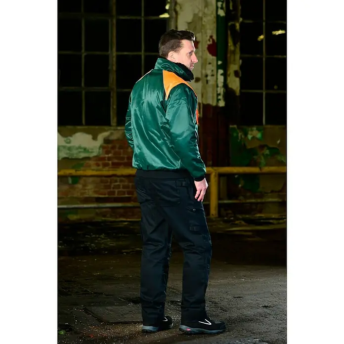 Forstblouson 4in1 - Grün/Orange, XXL - Der Mann trägt eine glänzend grüne Arbeitsjacke mit orangen Schultern und schwarze Arbeitshosen. Komplettiert wird der Look durch robuste Arbeitsschuhe.