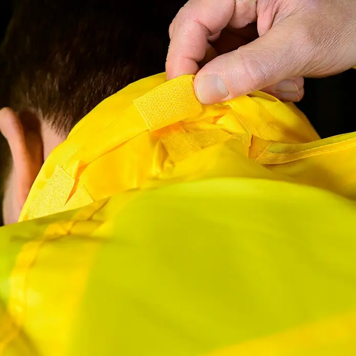 Regenjacke - M, 100% Polyester - Detailaufnahme eines gelben Kleidungsstücks mit Klettverschluss, das von einer Hand angepasst wird. Fokus auf Verschlussmechanismus.