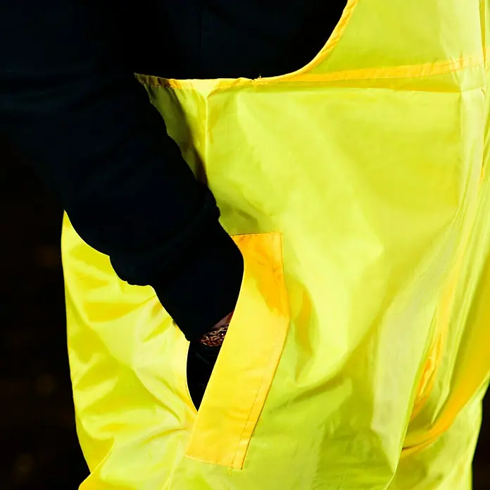 Regenlatzhose - M, 100% Polyester - Nahaufnahme einer gelben, wasserdichten Hose mit einer Hand in der Tasche, ideal für Arbeiten im Freien bei schlechtem Wetter.