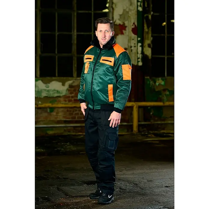 Forstblouson 4in1 - Grün/Orange, XXL - Eine Person trägt eine grüne Arbeitsjacke mit orangen Details und eine schwarze Arbeitshose. Der Fokus liegt auf der robusten Arbeitskleidung.