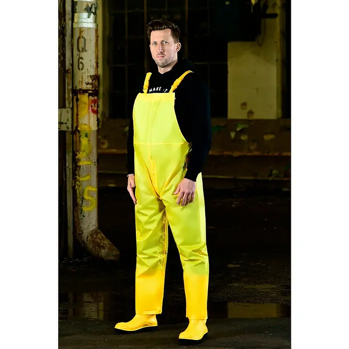 Regenlatzhose - M, Gelb, Kunststoff - Das Foto zeigt einen Mann in einer gelben wasserdichten Latzhose und Stiefeln, ideal für feuchte Umgebungen, vor einem industriellen Hintergrund.