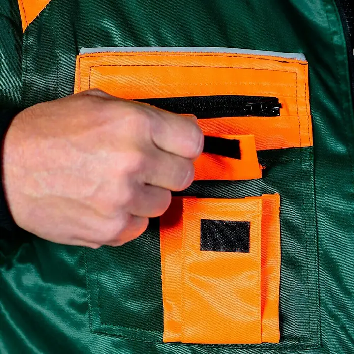 Forstblouson 4in1 - Grün/Orange, XXL - Detailansicht einer grünen Arbeitsjacke mit orangefarbenen Taschen und einer Hand, die eine Tasche öffnet. Praktische und robuste Arbeitskleidung.