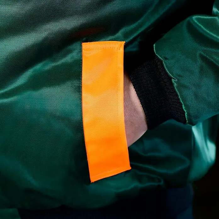 Forstblouson 4in1 - Grün/Orange, XXL - Detailaufnahme einer grünen Bomberjacke mit orangefarbenem Akzent. Die Jacke hat eine Seitentasche und ein geripptes Bündchen am Ärmel.
