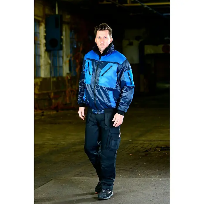 Elutex Piloten-Blouson 4in1 - Navy/Royalblau, XXL - Ein Mann in Arbeitskleidung, bestehend aus einer blauen Jacke und schwarzen Cargohosen, steht in einem Lagerhaus.