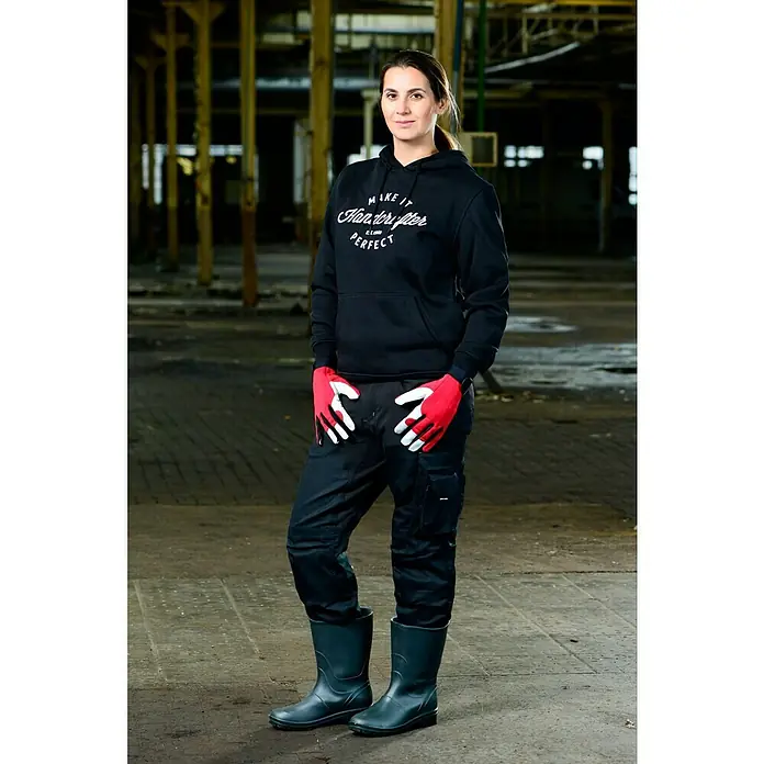 Guide Arbeitshandschuhe 54 PP - Rot/Weiß, 12 - Eine Frau in Arbeitskleidung, bestehend aus einem Hoodie, einer Hose, Stiefeln und Handschuhen, steht vor einem unscharfen Hintergrund.