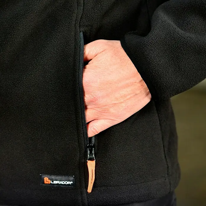 L.Brador Fleecejacke 687 P - Schwarz, 100 % Polyester, L - Nahaufnahme einer schwarzen Fleecejacke mit einer Hand in der Reißverschlusstasche, die die Textur und Details der Jacke hervorhebt.