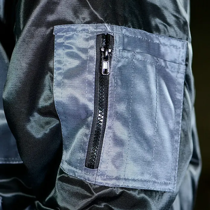 Elutex Piloten-Blouson 4in1 - Schwarz/Grau, M - Nahaufnahme einer grauen Jackentasche mit Reißverschluss. Der Reißverschluss ist schwarz und der Stoff der Tasche ist glänzend.