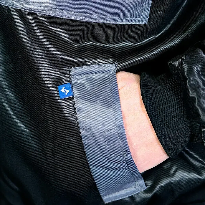 Elutex Piloten-Blouson 4in1 - Schwarz/Grau, M - Nahaufnahme einer Jackentasche mit einer Hand, die in die Tasche gesteckt ist. Die Jacke ist aus glänzendem, dunklem Material gefertigt.