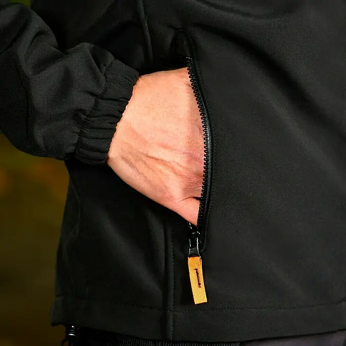 L.Brador Softshell-Jacke 554 P - Schwarz, L - Nahaufnahme einer schwarzen Jacke mit einer Hand in der Reißverschlusstasche.