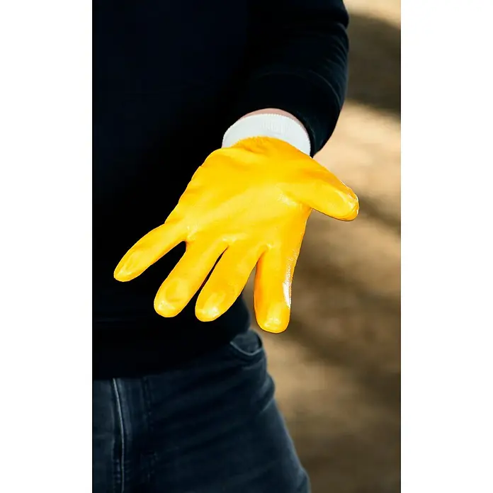 Guide Arbeitshandschuhe 807 PP - Gelb, 10 - Nahaufnahme einer gelben Gummihandschuh, getragen von einer Person in Jeans und einem schwarzen Pullover, der zum Schutz der Hände dient.