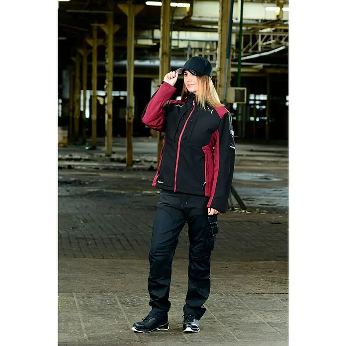 Flexfit Gorra - Negro, L/XL - Una mujer con ropa de trabajo, compuesta por una chaqueta negra y burdeos y unos pantalones, posa en una nave industrial.