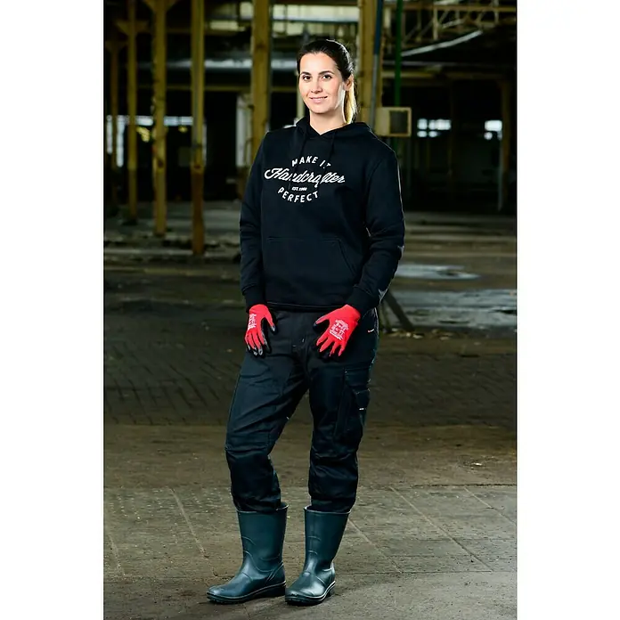 Wisent Arbeitshandschuhe Renovation - Schwarz/Rot, 8 - Eine Frau in Arbeitskleidung mit Handschuhen und Gummistiefeln vor einer Industriehalle, die Handwerkerkleidung präsentiert.