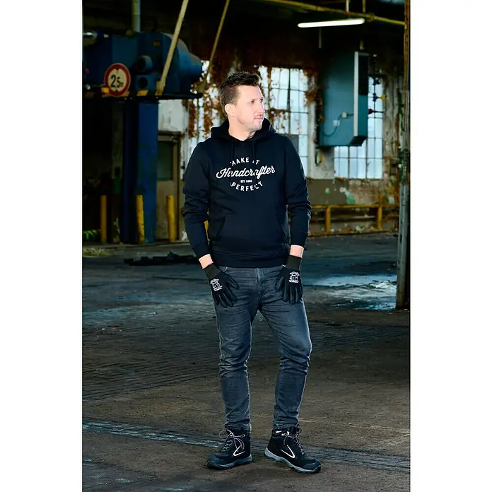 Wisent Arbeitshandschuhe Car-Repair - Schwarz, 8 - Ein Mann in schwarzem Hoodie mit Aufschrift und Arbeitshandschuhen und Sicherheitsschuhen in einer Industriehalle.