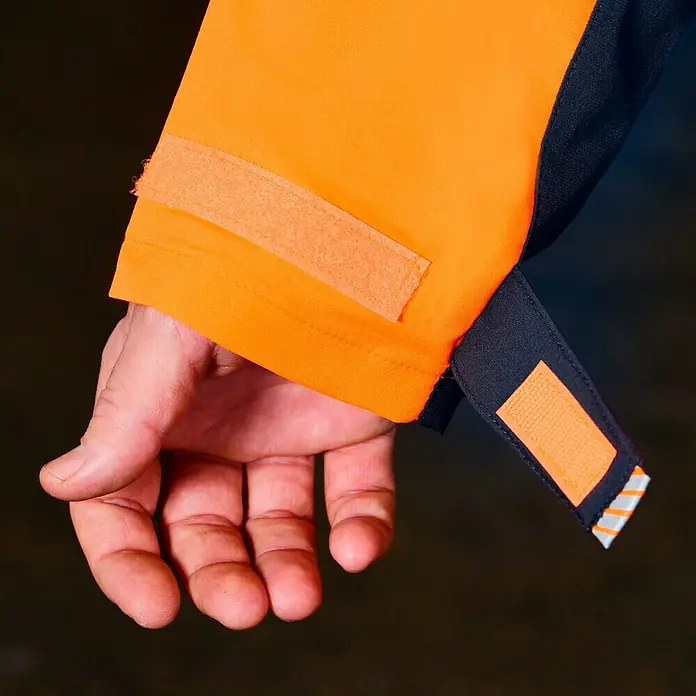 Bullstar Multifunktionsjacke Proximo - Orange/Anthrazit, L - Detailaufnahme einer orangefarbenen Jacke mit Klettverschluss und einer Hand, die die Jacke hält.