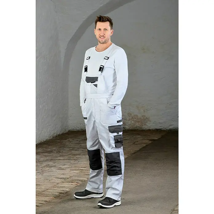 Uvex Sicherheitshalbschuh 1 Sport - 42, Schutzklasse nach EN ISO 20345: S1 - Ein Mann in weißer Latzhose mit grauen Akzenten und einem weißen Langarmshirt steht vor einer Wand. Die Latzhose hat mehrere Taschen.