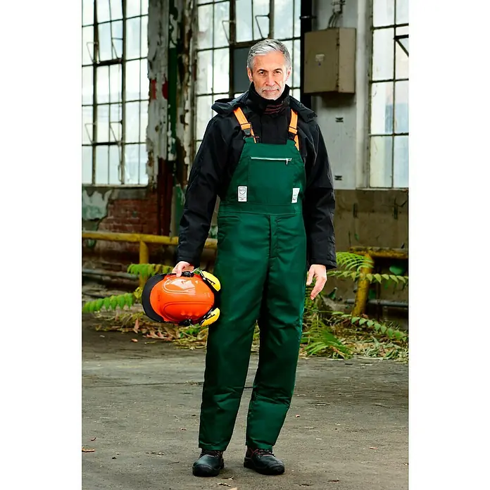 Bullstar Forst-Schutzhelm Peltor Master - Orange - Mann in grüner Arbeitskleidung hält einen Helm in einem Lager. Robuste grüne Arbeitskleidung mit orangefarbenen Trägern.