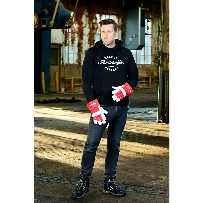 Wisent Arbeitshandschuhe Vechta - Handschuhgröße: 10, Natur/Rot - Mann in schwarzem Hoodie mit Aufschrift und Arbeitshandschuhen, der in einer Fabrikhalle steht. Ideal für Handwerker und Arbeiter.
