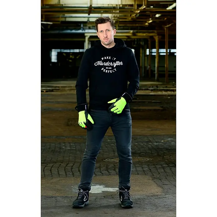 Guide Arbeitshandschuhe 5148W - 9 - Ein Mann in Arbeitskleidung mit Handschuhen und Stiefeln steht in einer Fabrikhalle. Die Kleidung ist schwarz mit auffälligen gelben Akzenten.