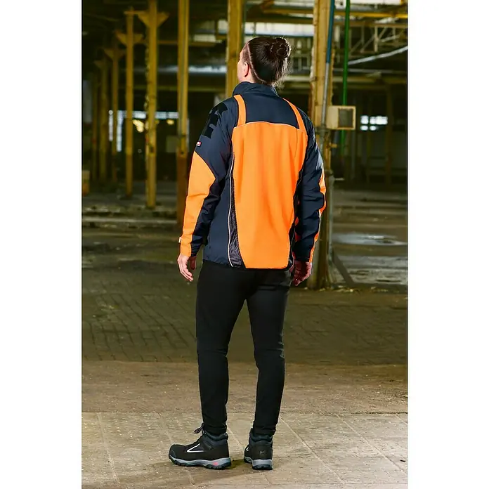 Bullstar Multifunktionsjacke Proximo - Orange/Anthrazit, L - Eine Person trägt eine orange-marineblaue Arbeitsjacke und schwarze Hosen mit Stiefeln in einer Industrieumgebung.