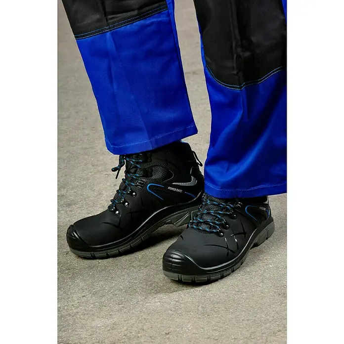 Power Safe Sicherheitshalbschuh Tom - Schuhgröße: 46, S3 - Nahaufnahme von schwarzen Power Safe-Sicherheitsstiefeln mit blauen Akzenten, getragen mit blauen und schwarzen Arbeitshosen auf grauem Grund.
