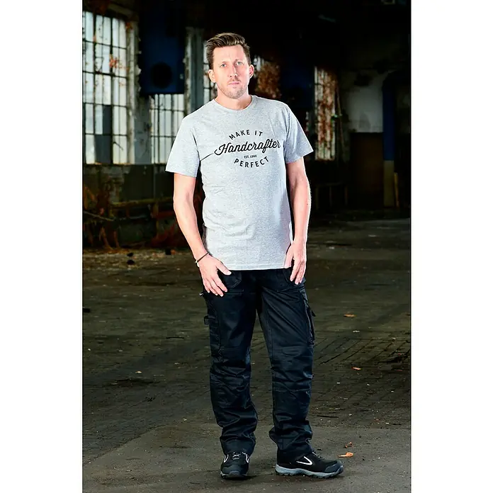 T-Shirt Handcrafter - Grau, L - Ein Mann steht in Arbeitskleidung mit einem grauen T-Shirt mit Aufdruck und schwarzen Cargo-Hosen. Ideal für Handwerker und den Arbeitsalltag.