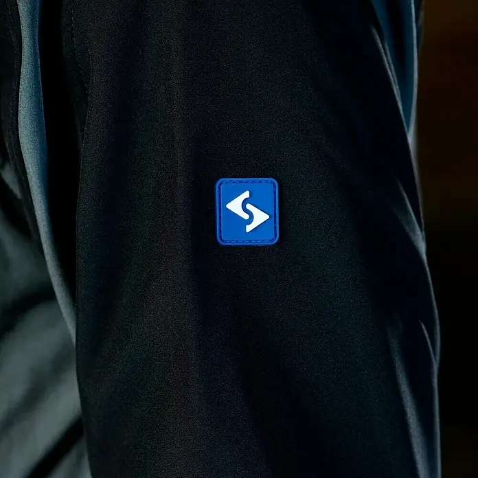 HD Concept Softshell-Jacke Fit - Schwarz/Grau, XXL - Detailaufnahme einer schwarzen Jacke mit blauem, quadratischem Logo-Patch auf dem Ärmel, das ein weißes Symbol zeigt.