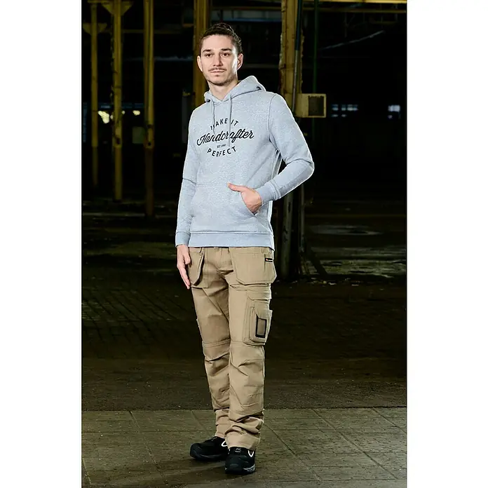 Kapuzenpullover Handcrafter - Grau, Größe: XXXL - Ein Mann in Handwerkerhose und grauem Kapuzenpullover mit Aufdruck steht in einer Fabrikhalle.