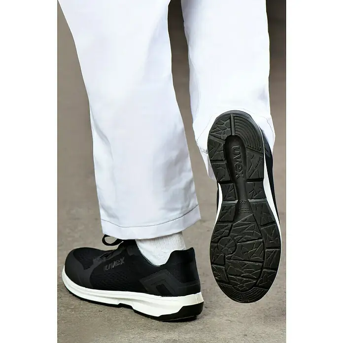 Uvex Sicherheitshalbschuh 1 Sport - 44, Schutzklasse nach EN ISO 20345: S1 - Das Bild zeigt einen Uvex-Sicherheitsschuh mit schwarzem Obermaterial und weißer Sohle, der getragen wird. Die Sohle des Schuhs ist gut sichtbar.