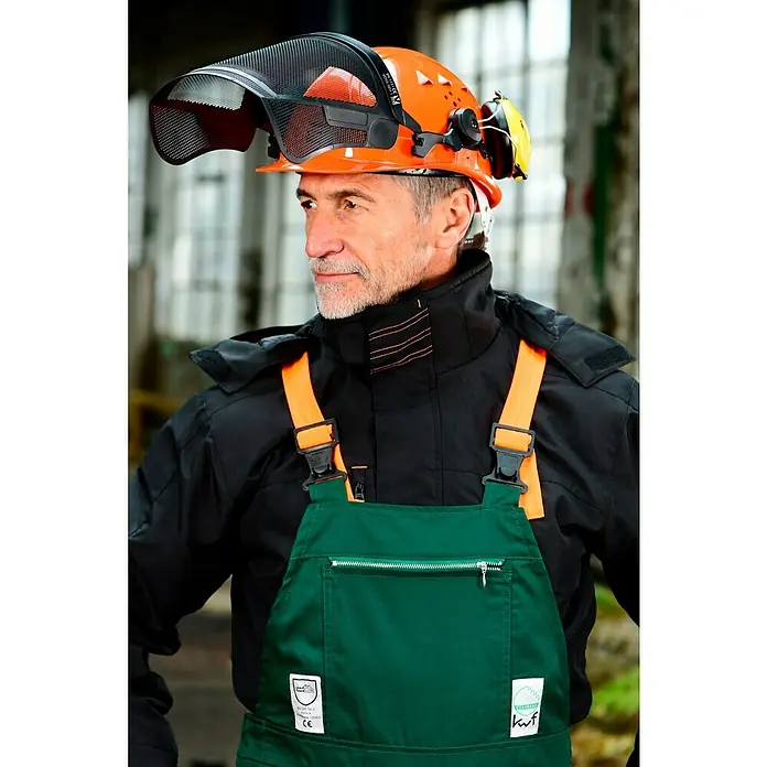 Bullstar Forst-Schutzhelm Peltor Master - Orange - Ein Mann trägt eine Schutzausrüstung inklusive Helm mit Visier und Gehörschutz sowie eine grüne Latzhose mit schwarzer Jacke für Forstarbeiten.