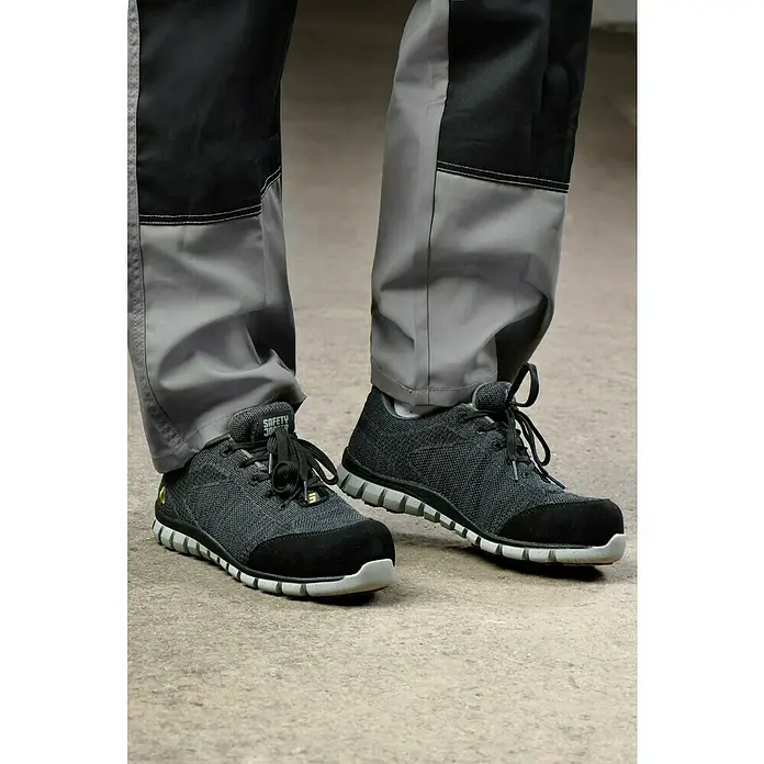 Safety Jogger Sicherheitshalbschuh Morris S1P - 45, Schutzklasse nach EN ISO 20345: S1P, Schwarz - Nahaufnahme von Sicherheitsschuhen Safety Jogger mit grauer Textur und schwarzen Details, getragen mit grauen Arbeitshosen.