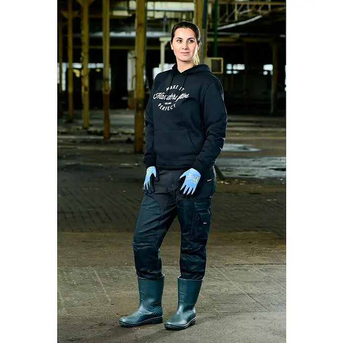 OX-ON Arbeitshandschuhe Recycle Basic 16002 - Blau, 8 - Eine Frau in Arbeitskleidung, bestehend aus einem schwarzen Hoodie, einer Arbeitshose und Gummistiefeln, steht in einer Fabrikhalle.