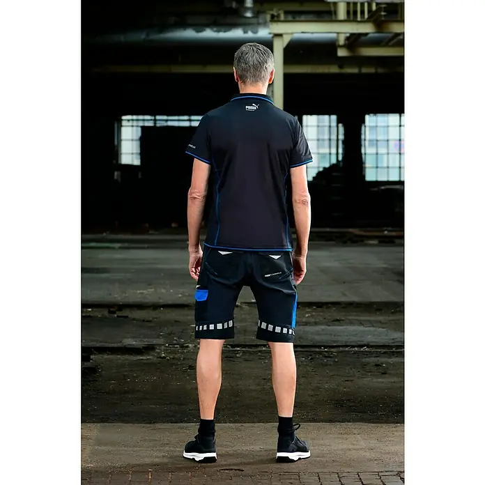Puma Workwear Poloshirt Champ - Carbon, XL - Rückenansicht einer Person, die ein Puma-Polohemd und Arbeitsshorts mit reflektierenden Details sowie schwarze Schuhe trägt.