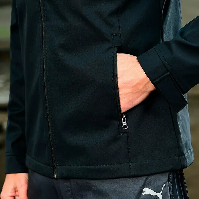 Puma Workwear Softshell-Jacke Champ - Carbon/Blau, M - Detailaufnahme einer schwarzen Jacke mit Reißverschluss und einer Hand in der Tasche. Sportliche Eleganz für jeden Tag.