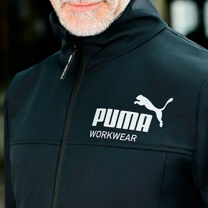 Puma Workwear Softshell-Jacke Champ - Carbon/Blau, M - Schwarze Puma-Arbeitsjacke mit weißem Logo für Komfort und Stil bei der Arbeit.