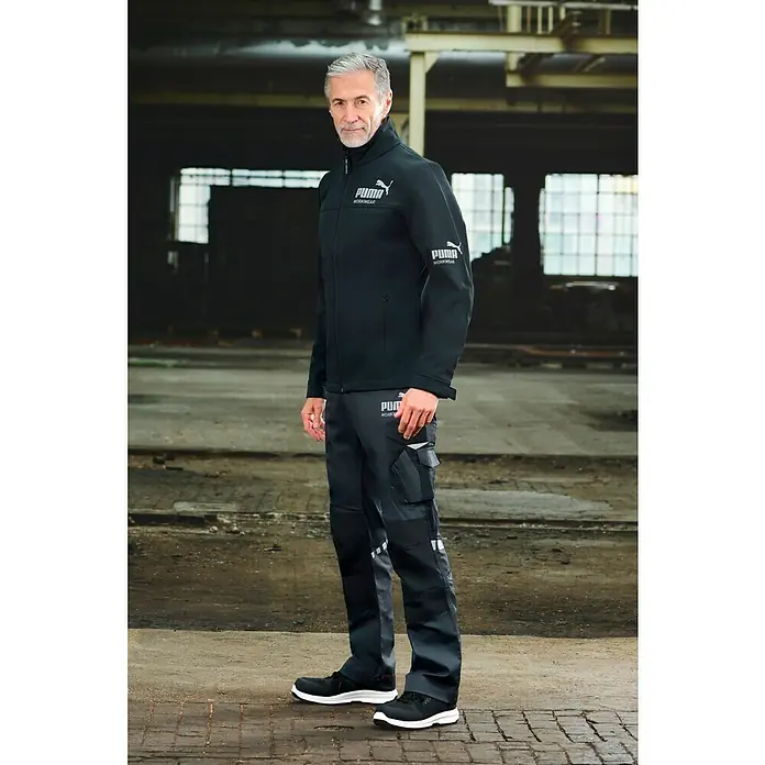 Puma Workwear Softshell-Jacke Champ - Carbon/Blau, M - Der Mann trägt eine schwarze Puma-Arbeitsjacke und passende Hose. Das Outfit wird durch Sicherheitsschuhe vervollständigt.