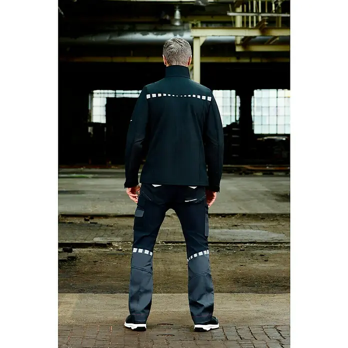 Puma Workwear Softshell-Jacke Champ - Carbon/Blau, M - Eine Person von hinten in Arbeitskleidung. Die Jacke ist schwarz mit Reflektoren. Die Arbeitshose ist schwarz mit grauen Partien und Reflektoren.