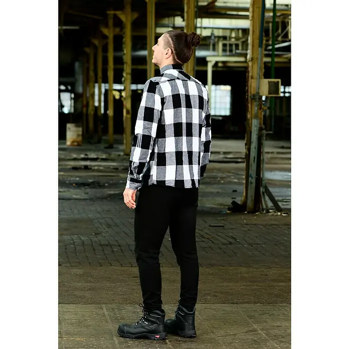 Brandit Flanellhemd - Schwarz/Weiß, XXXL - Ein Mann mit einem Haarknoten, der ein schwarz-weiß kariertes Hemd, eine schwarze Hose und schwarze Stiefel in einem Lagerhaus trägt.