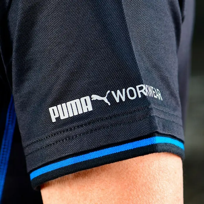 Puma Workwear Poloshirt Champ - Carbon, XL - Nahaufnahme eines dunkelblauen Puma Workwear-Shirts mit blauem Detail am Ärmel. Das Puma Logo ist in Weiß aufgedruckt.