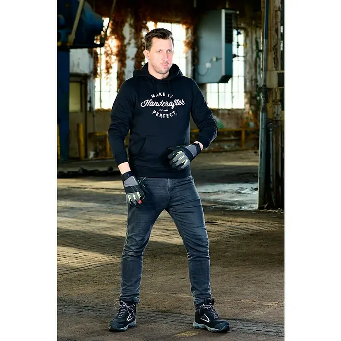Guide Arbeitshandschuhe 5001 - Schwarz, 10 - Der Mann mit dem schwarzen Hoodie "Make it Handcrafter Perfect" und Arbeitshandschuhen steht selbstbewusst in einer alten Industriehalle.
