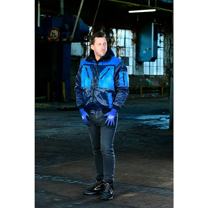 Guide Arbeitshandschuhe 578 HP - Dunkelblau, 6 - Der Mann trägt eine stylische, blau-schwarze Bomberjacke, blaue Handschuhe, graue Jeans und schwarze Stiefel in einer Industriehalle.